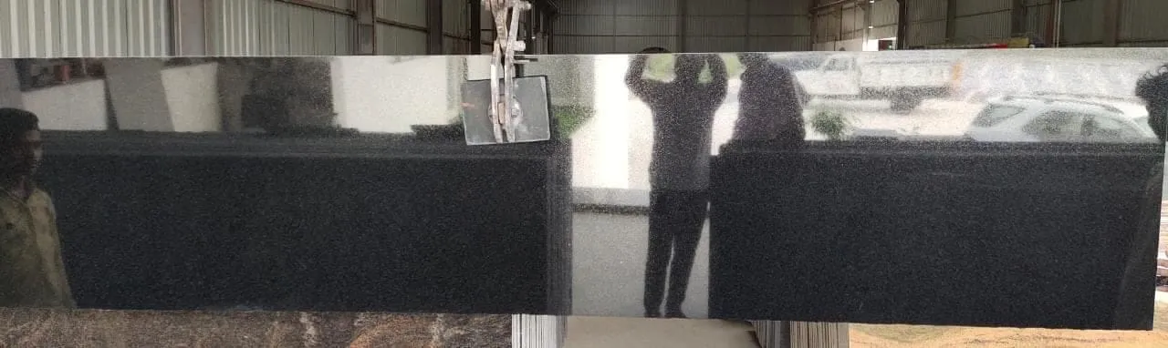 A large, polished black granite slab is held upright inside an industrial facility, reflecting people and objects in its shiny surface.