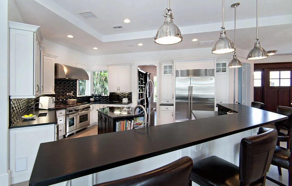 Modern kitchen with black countertops, stainless steel appliances, white cabinets, pendant lights, and bar seating. A window above the sink lets in natural light.